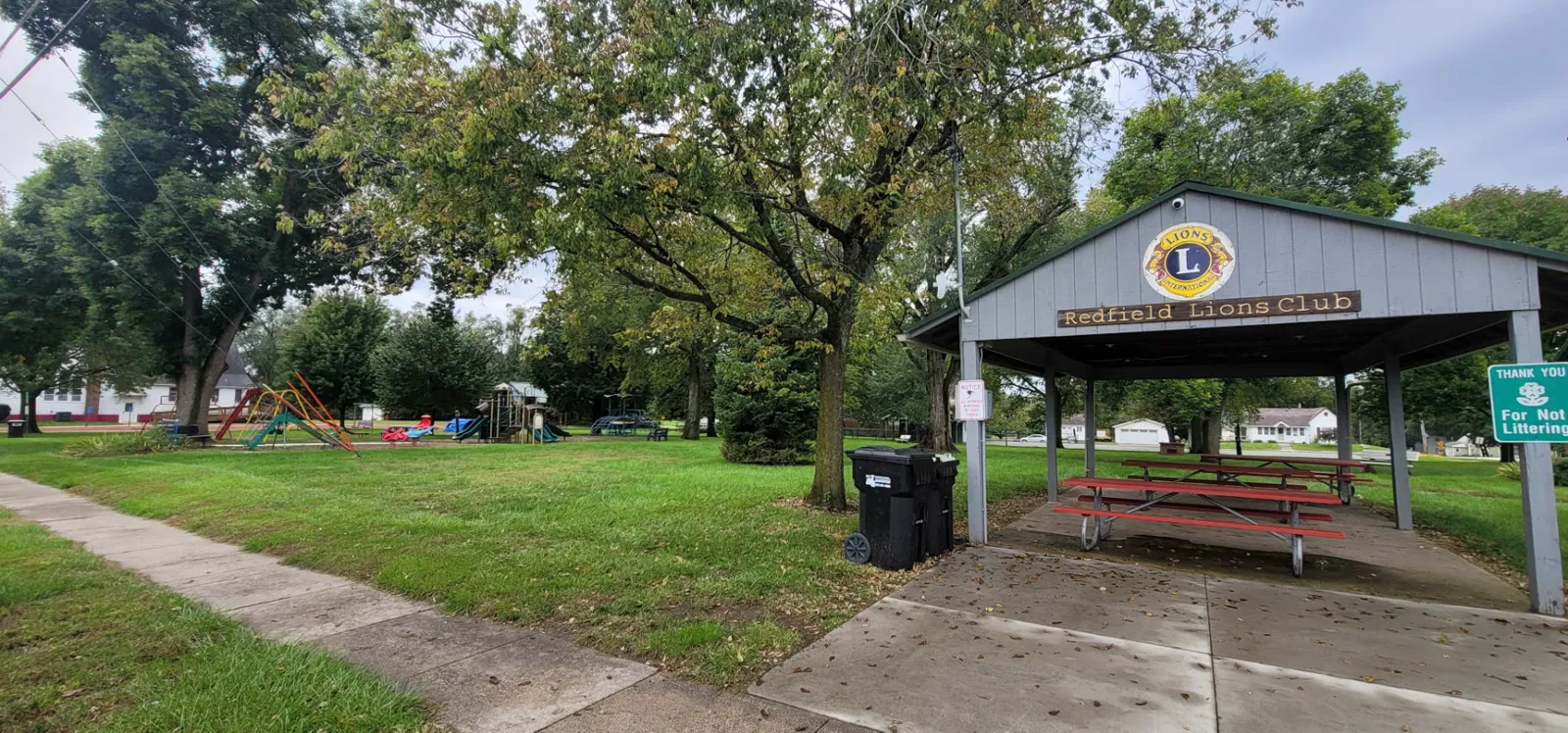Redfield City Park's playground and picnic shelter seen across the open lawn beneath a large shade tree