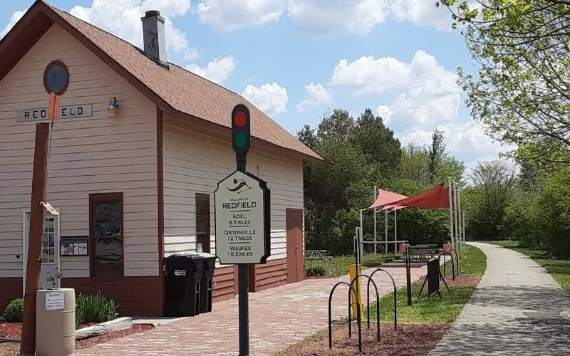 The restored Redfield depot trailhead with a welcome sign listing trail distances, bike racks, and the paved Raccoon River Valley Trail heading east