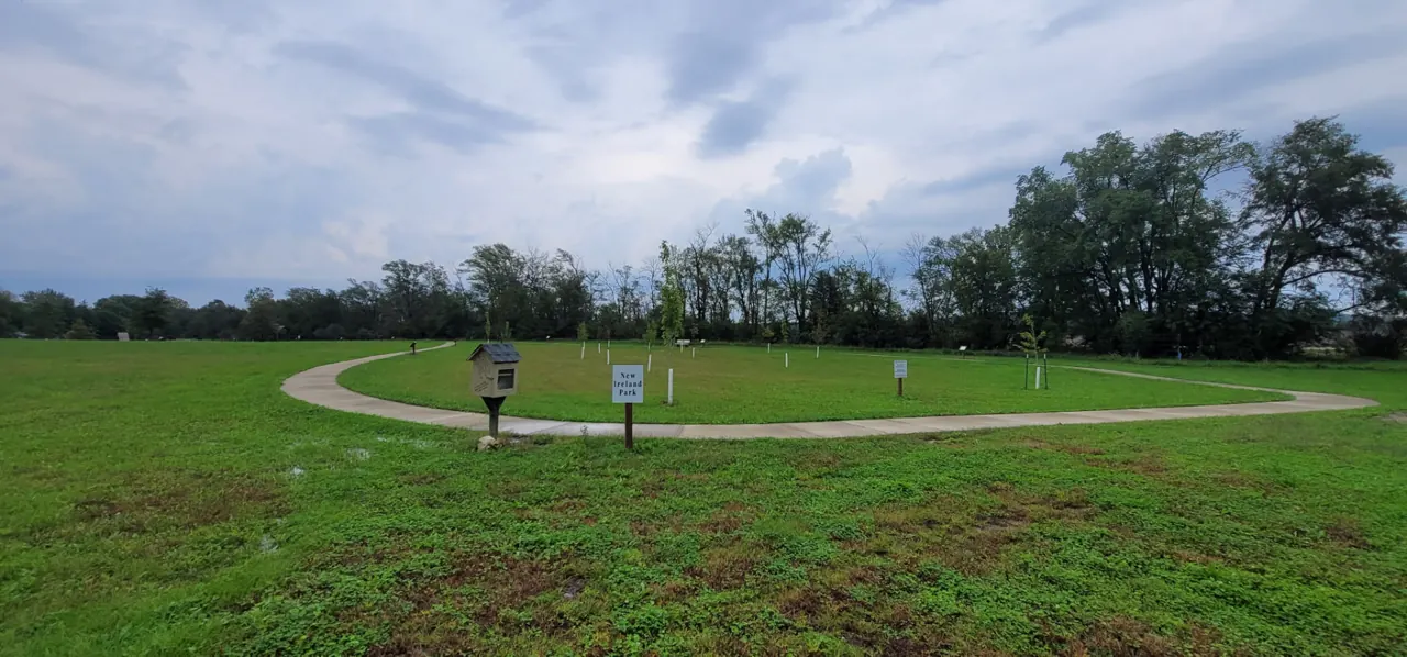 The paved walking path at New Ireland Park curving past the park's white entry sign and a little free library, with young saplings staked along the lawn and a dense row of mature trees beneath an overcast sky