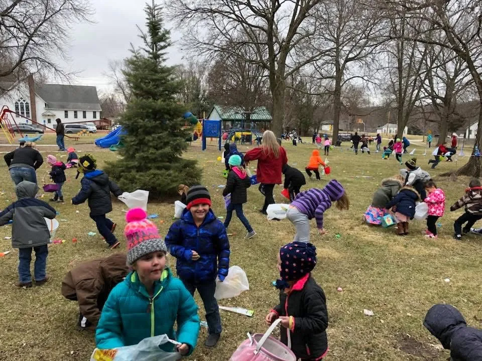 Children in winter hats and coats collecting eggs across the park lawn near the playground