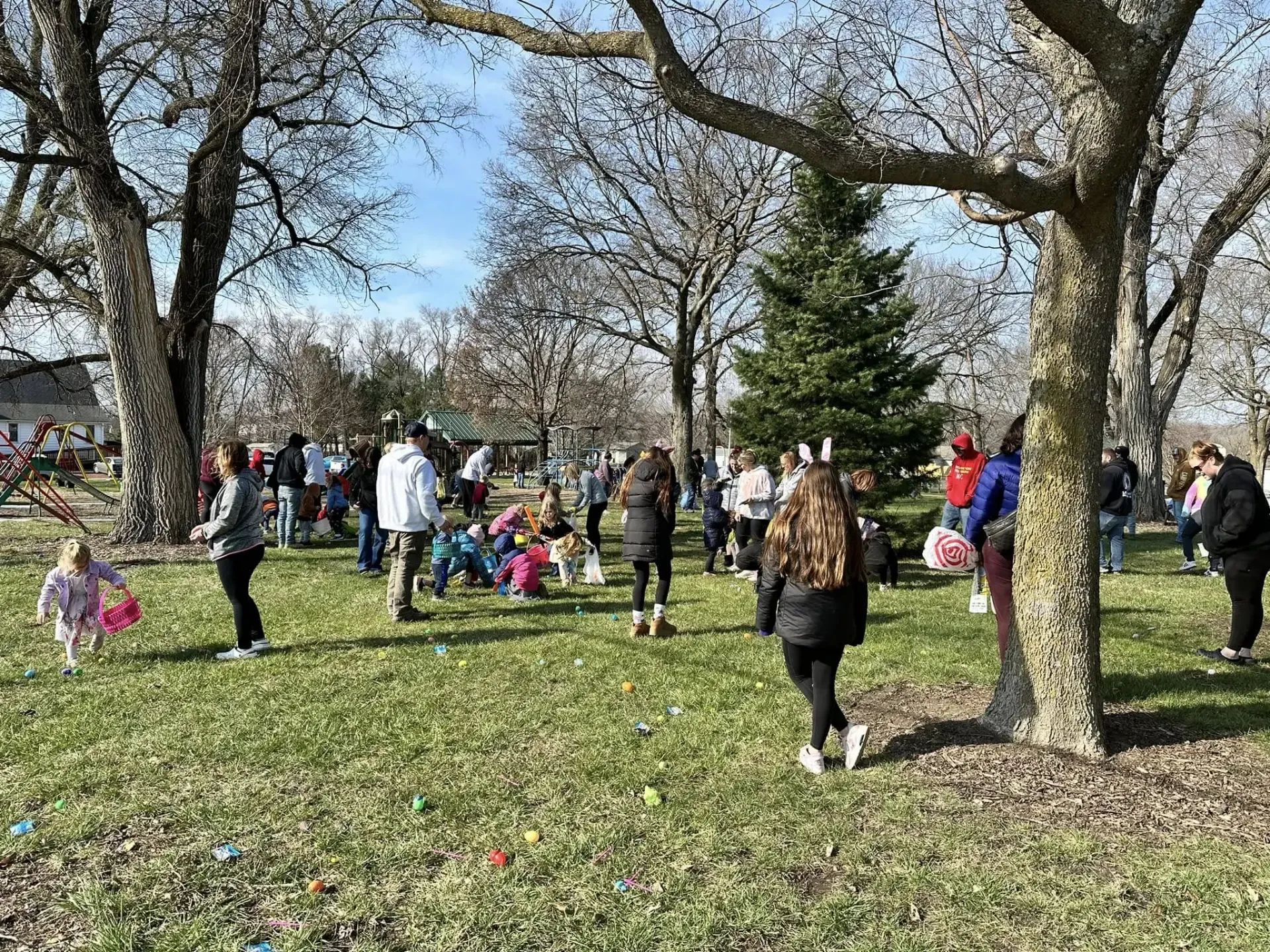 Kids and families searching for colorful plastic eggs under tall trees at Redfield City Park during the Easter Egg Hunt