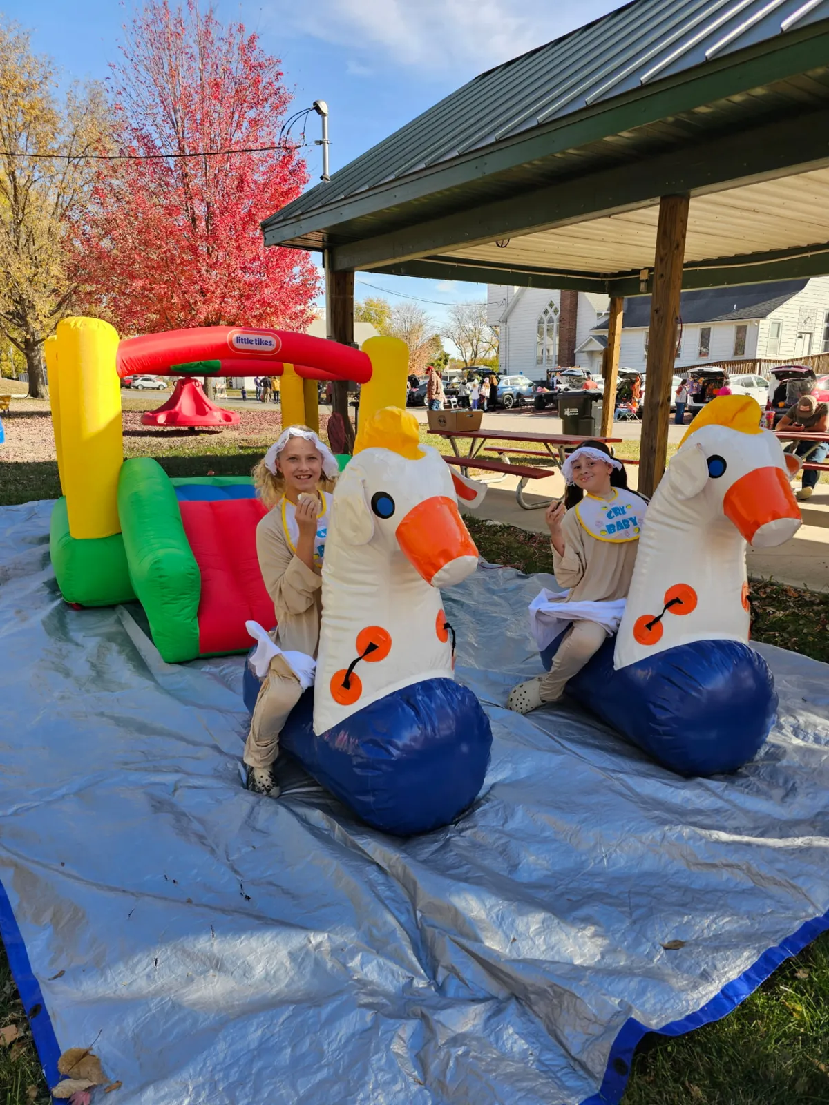 Two kids in matching cry-baby costumes riding inflatable ponies beside a bounce house under red fall leaves at the park shelter