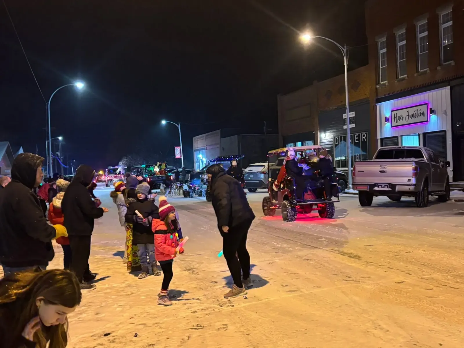 Families with glow sticks lining a snowy downtown Redfield street at night as lighted vehicles roll by in the Winter Nights Lighted Parade